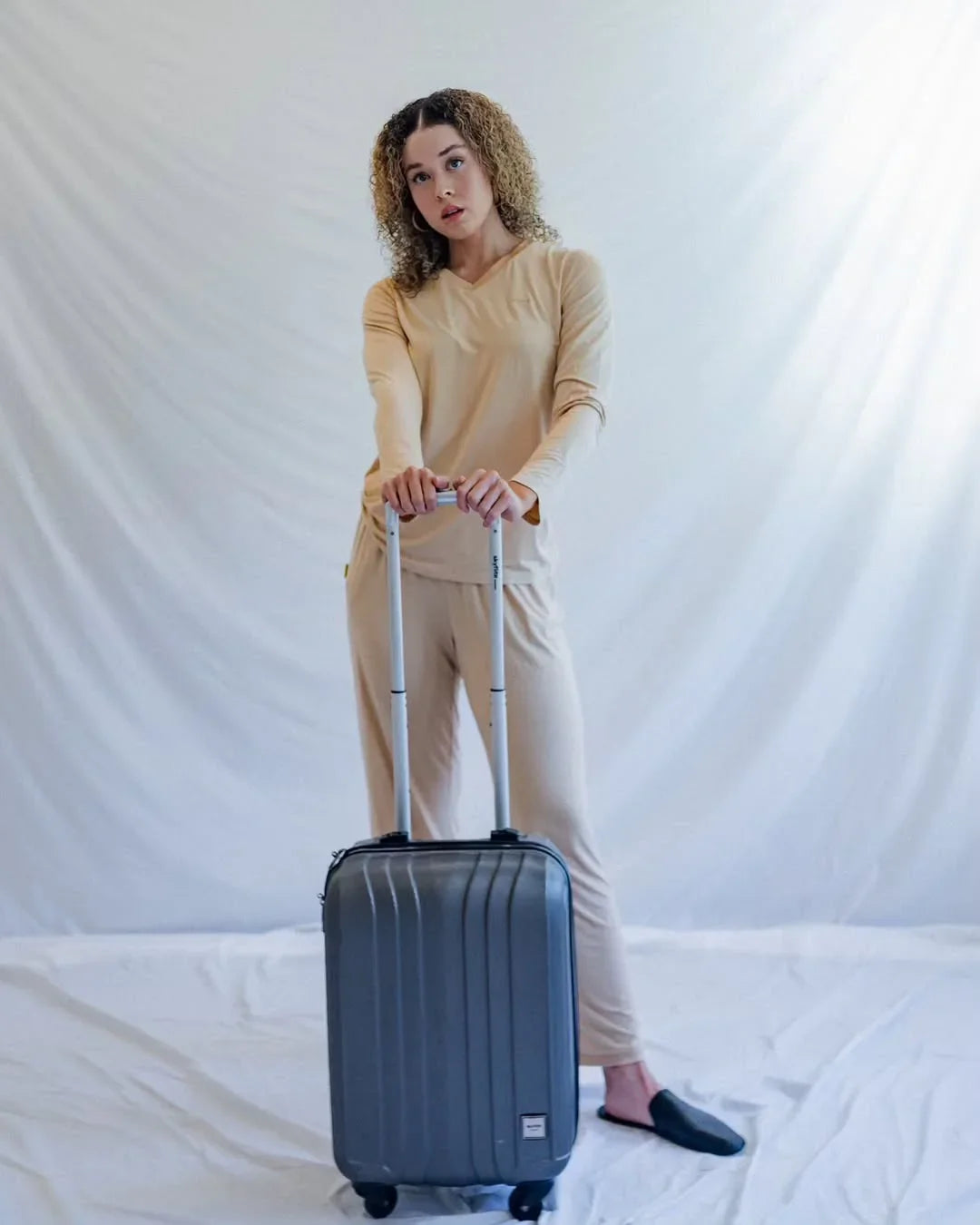 Woman in beige loungewear with a silver carry-on suitcase standing on white background