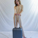 Woman in beige loungewear with a silver carry-on suitcase standing on white background