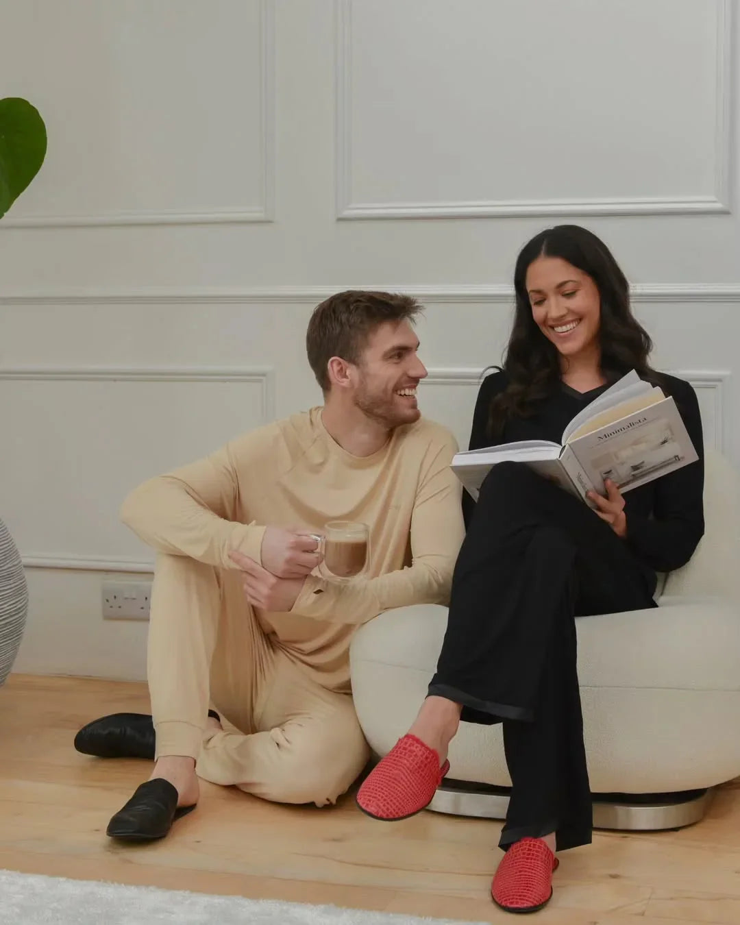 Smiling couple relaxing indoors, man in beige loungewear with coffee, woman reading in black outfit and red slippers