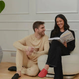 Smiling couple relaxing indoors, man in beige loungewear with coffee, woman reading in black outfit and red slippers
