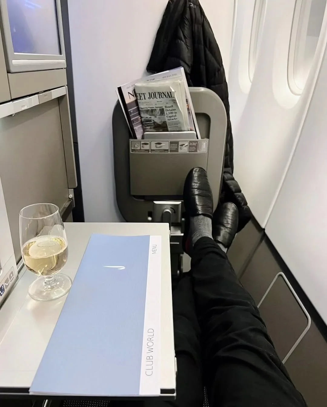 Business class airplane seat with legs up, Club World menu, wine glass, and newspaper