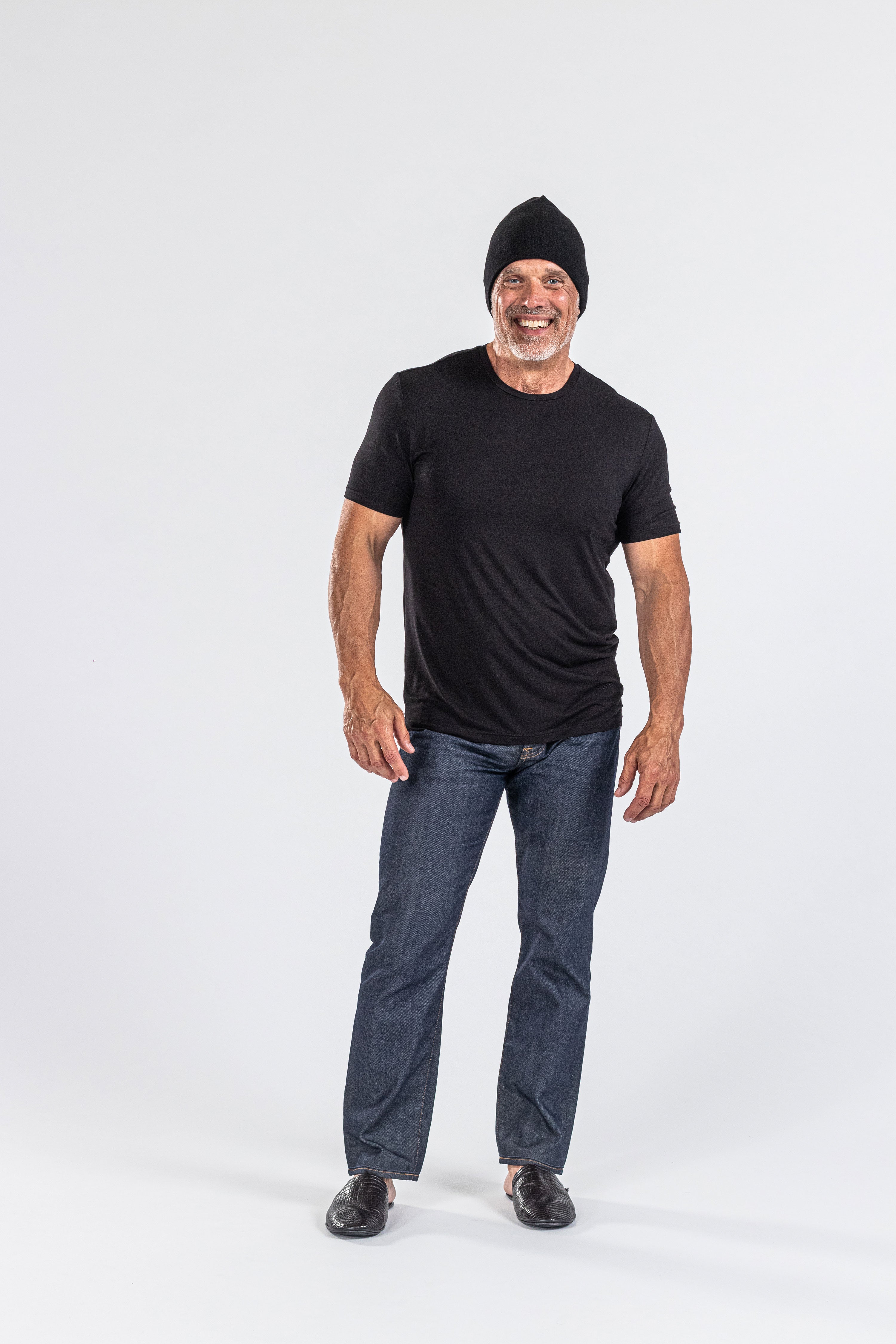 Smiling man wearing a black cashmere hat, black t-shirt, jeans, and loafers, studio backdrop