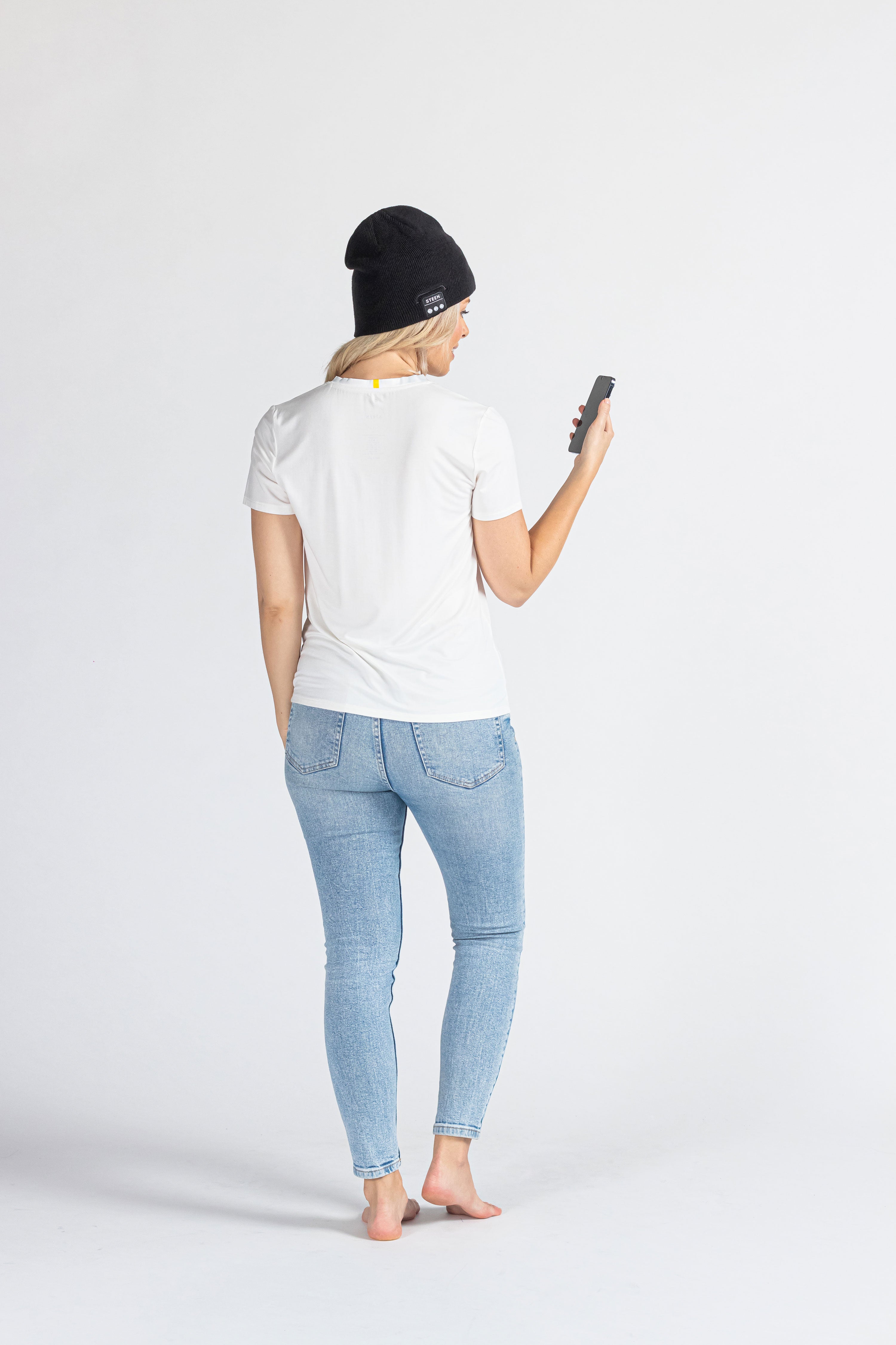 Woman wearing black music beanie, white t-shirt, jeans, holding phone, studio background