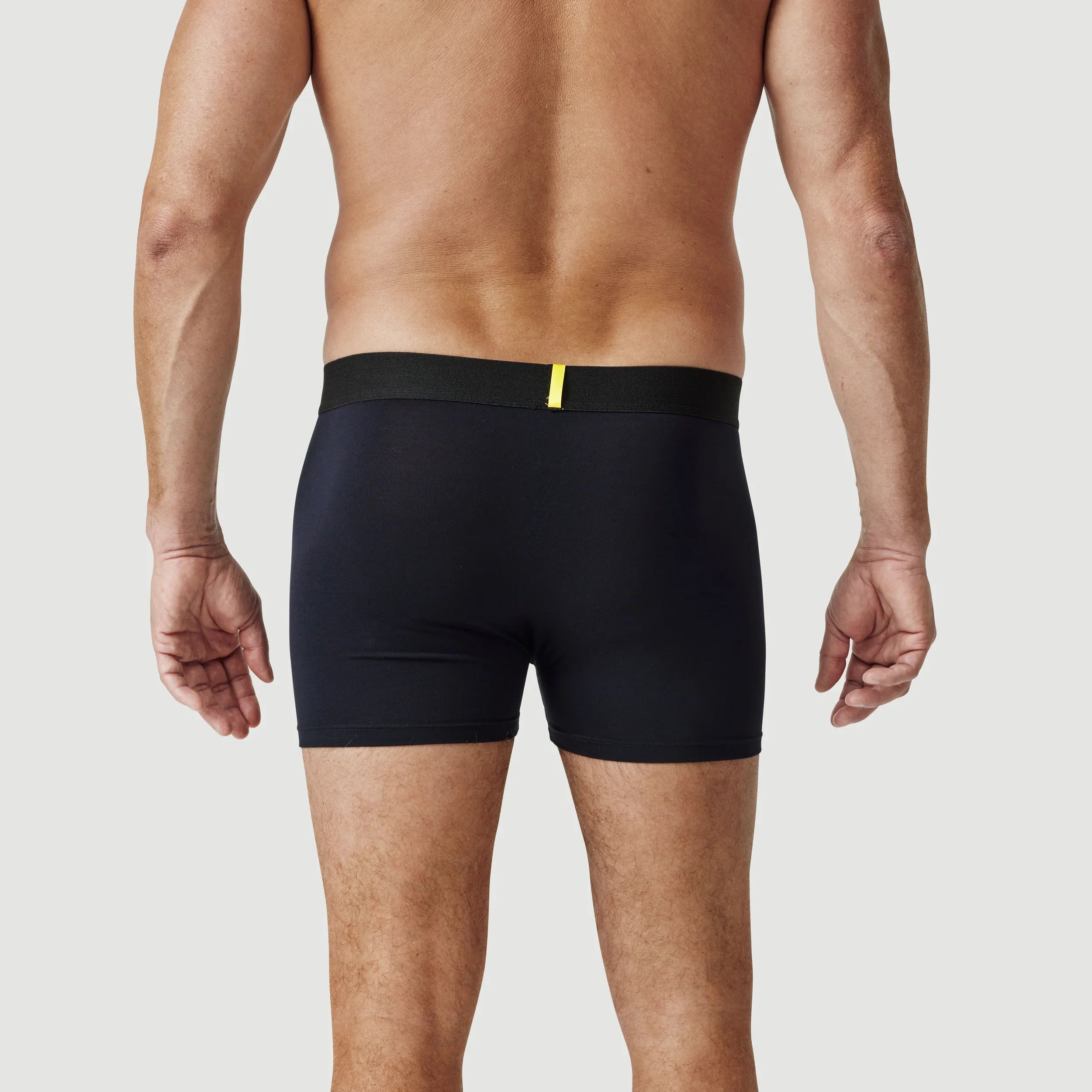 Man wearing black boxer briefs with yellow waistband detail, seen from the back on white background