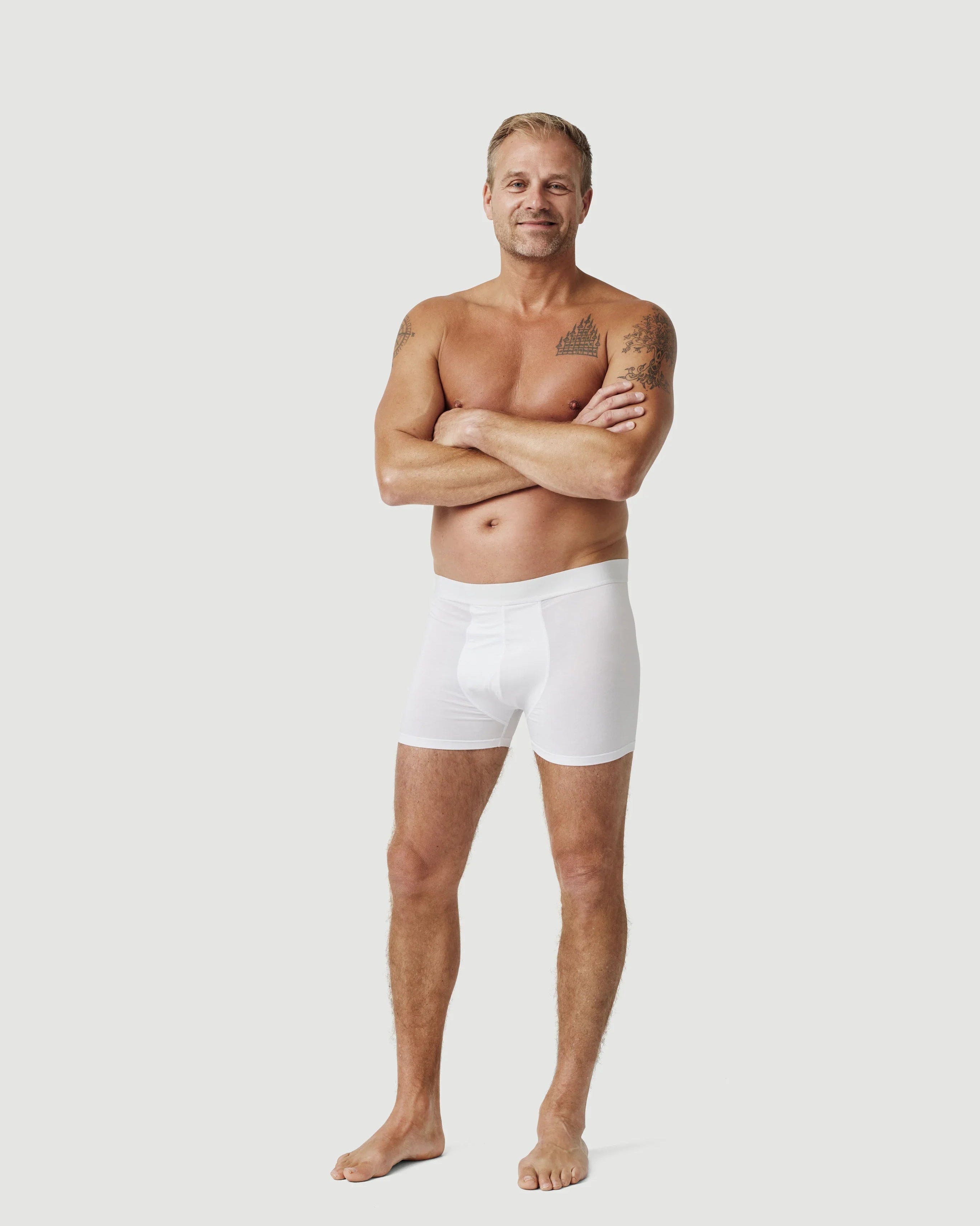Smiling man with tattoos modeling white boxer briefs on plain background