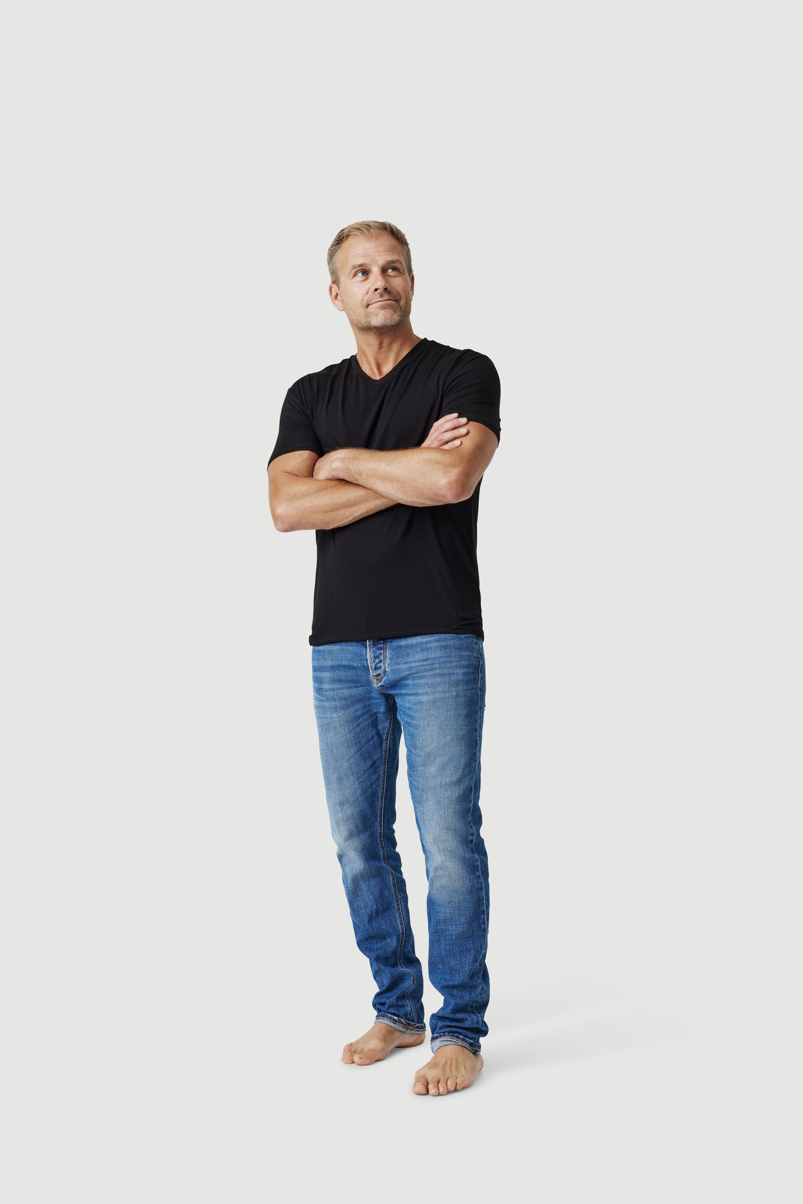 Man in black t-shirt and blue jeans standing barefoot against white background