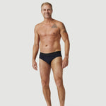 Smiling man wearing black men's swim briefs against a plain white background