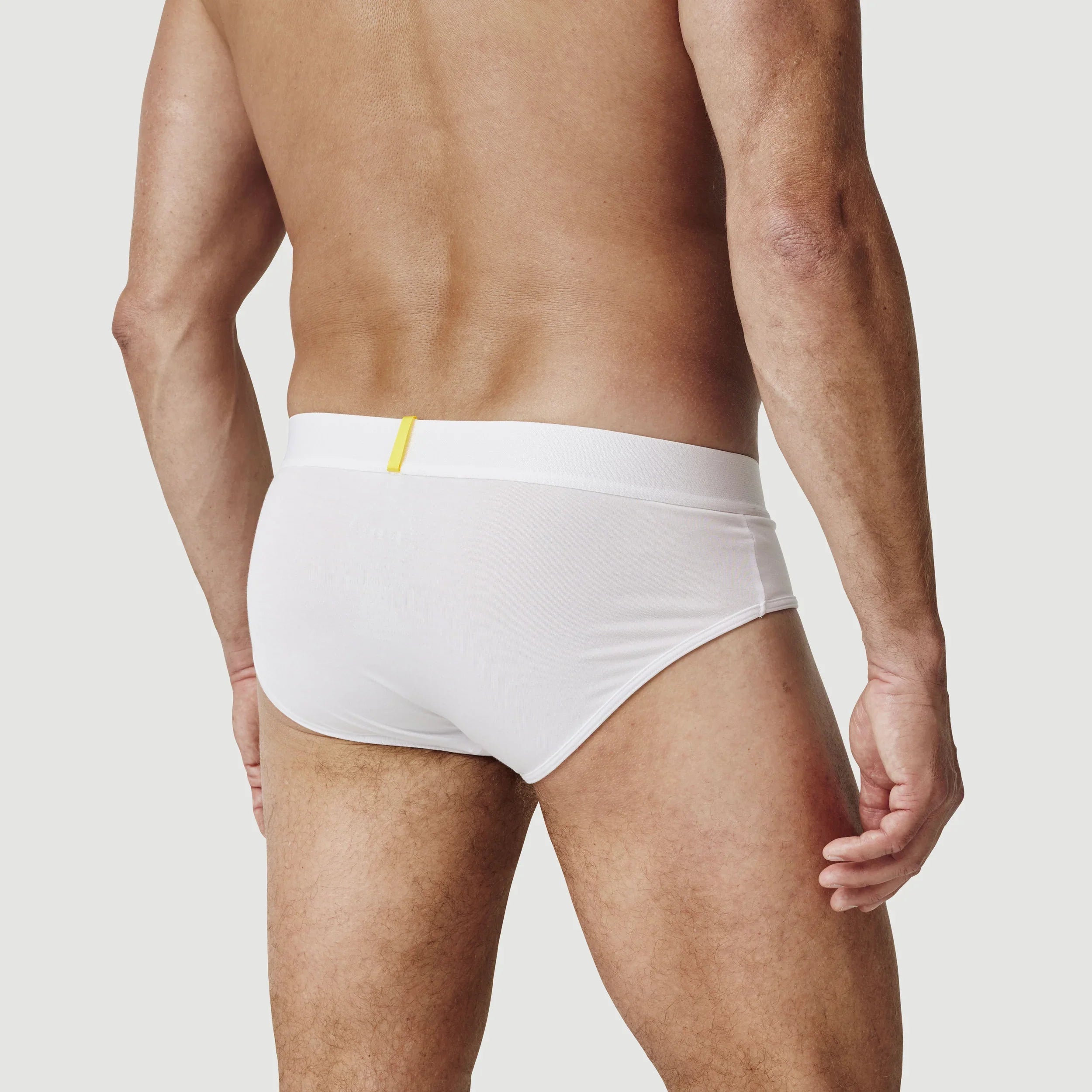 Man wearing white briefs underwear with yellow tag, shown from the back against plain background