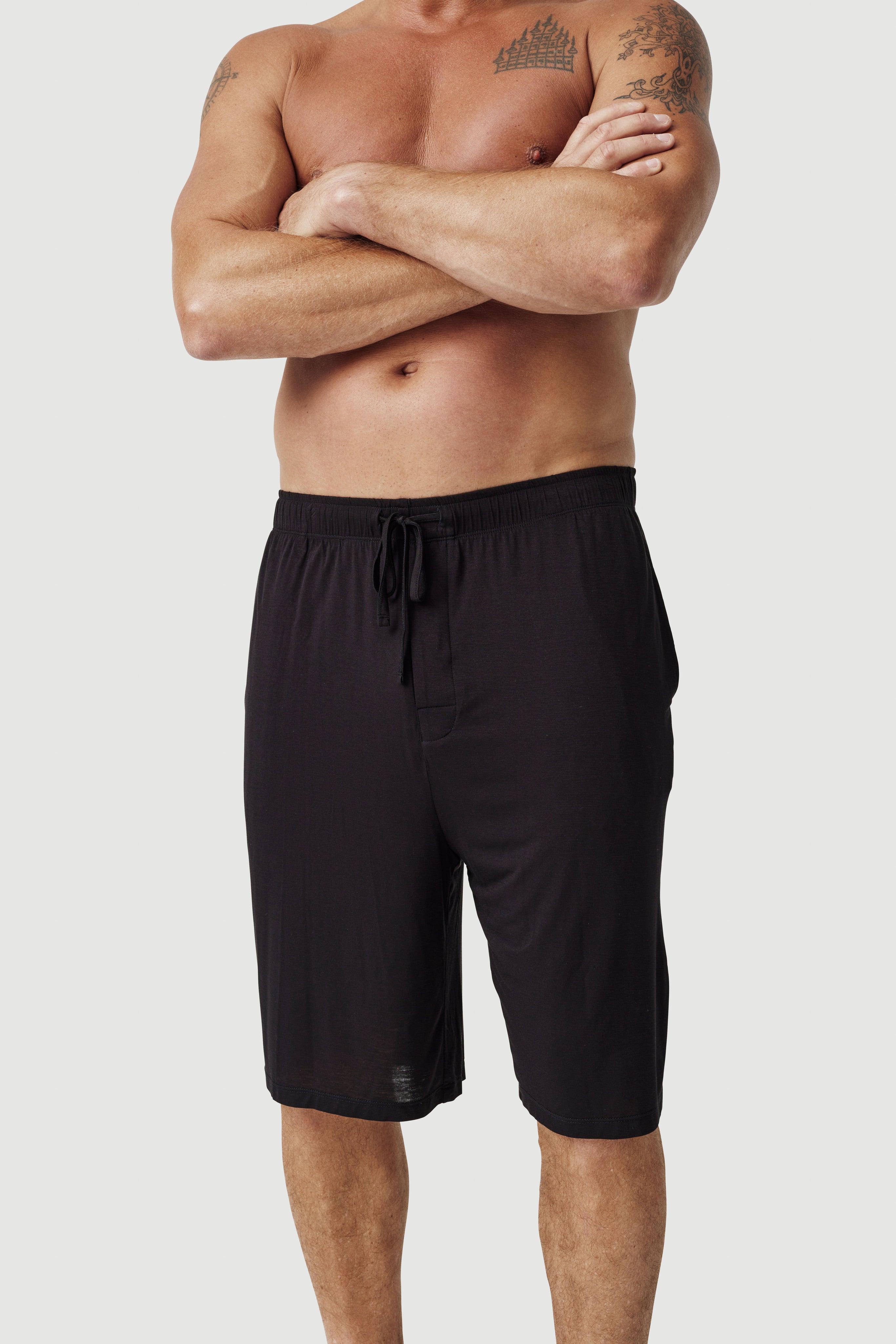 Man wearing black drawstring lounge shorts on white background, tattoos visible