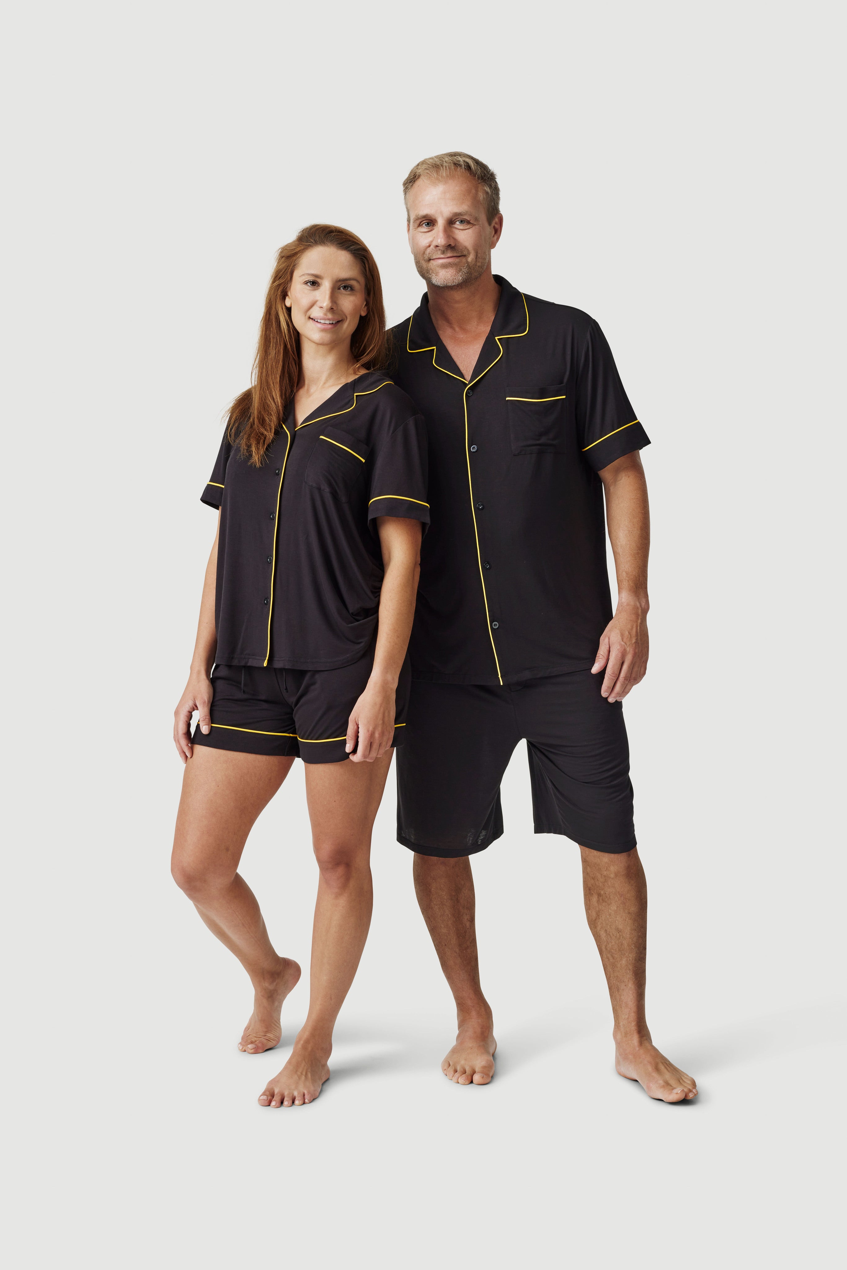 Man and woman wearing matching black short sleeve pajamas with yellow piping, on white background
