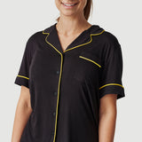 Woman wearing black short sleeve pajama top with yellow piping, smiling, on light background.