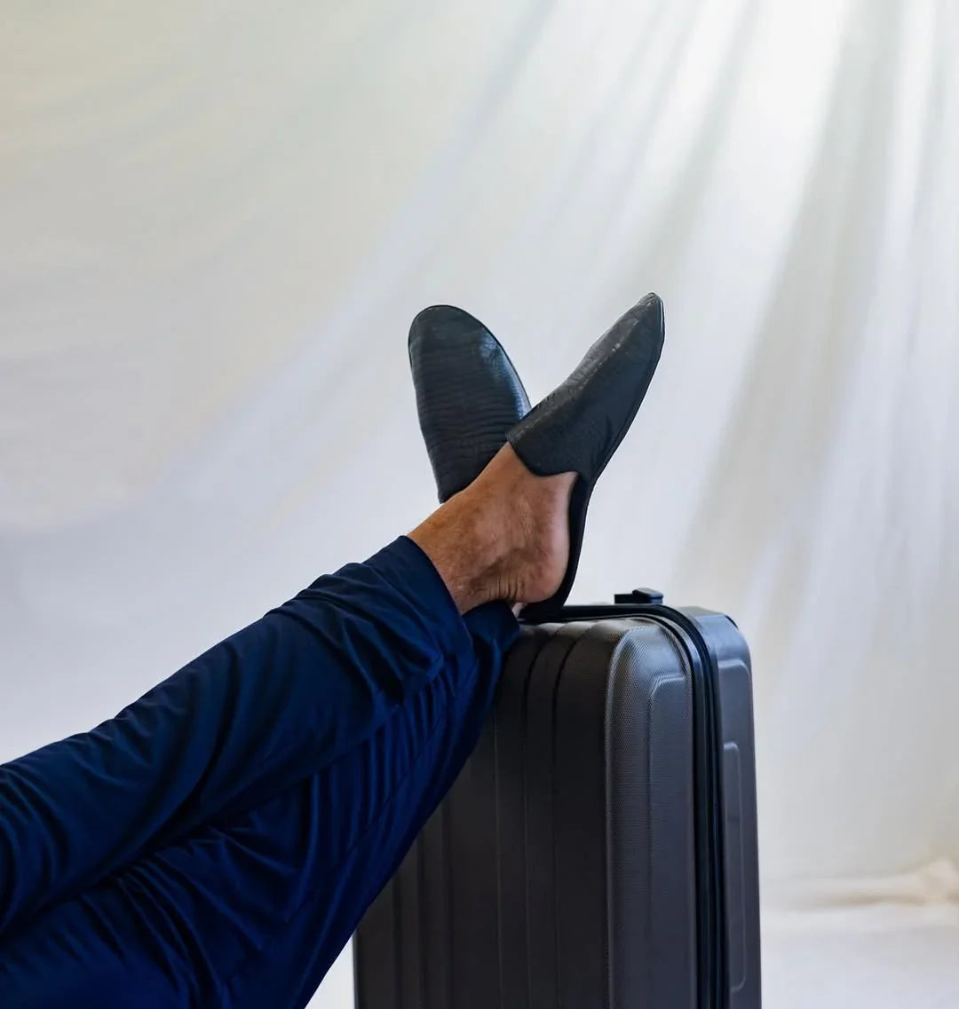 Man in navy pants wearing black leather travel slippers, feet on a suitcase, white backdrop