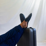 Man in navy pants wearing black leather travel slippers, feet on a suitcase, white backdrop