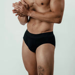Man wearing black bamboo briefs, standing side view, showing elastic waistband and leg tattoo
