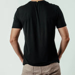 Man wearing black short sleeve t-shirt, beige pants, shown from the back, plain background