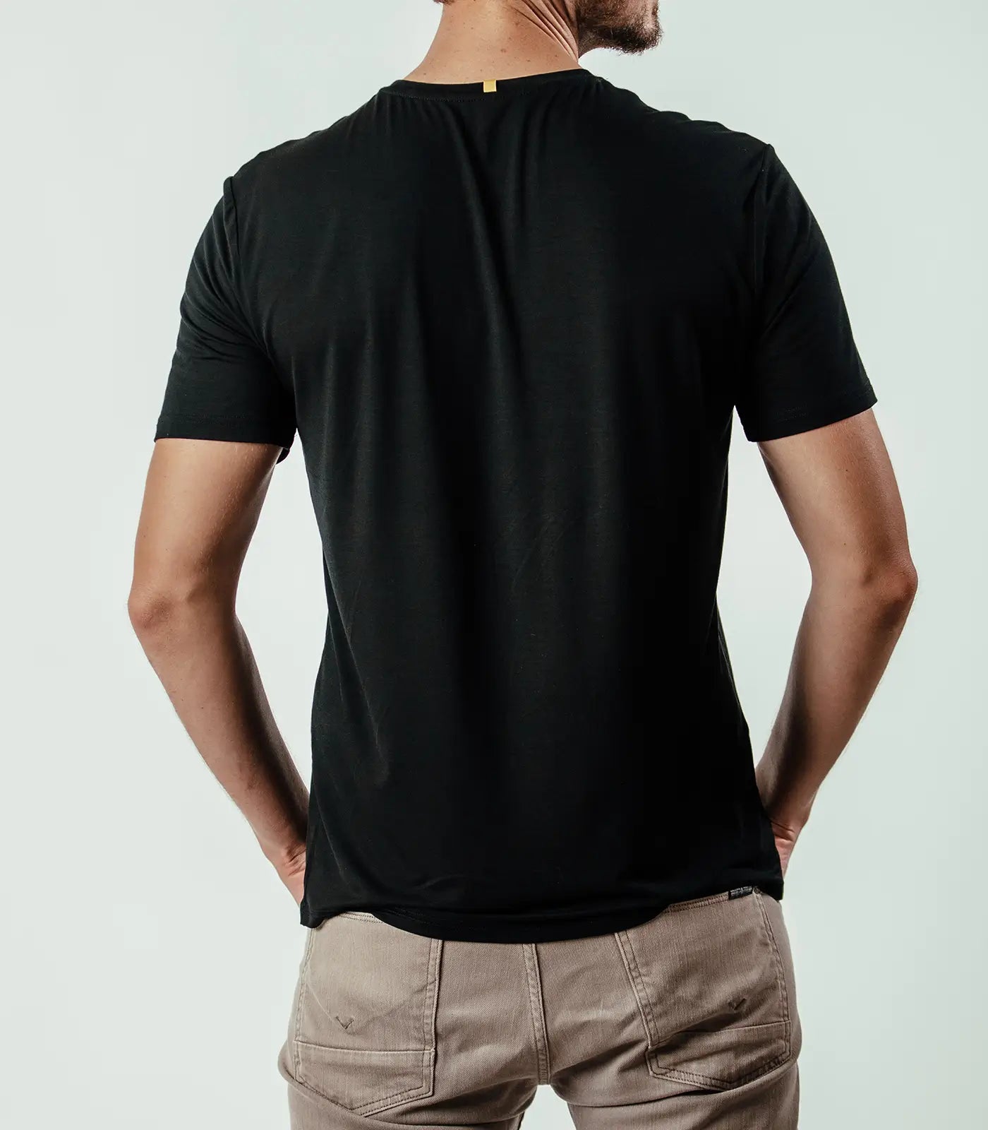 Man wearing black short sleeve t-shirt, beige pants, shown from the back, plain background